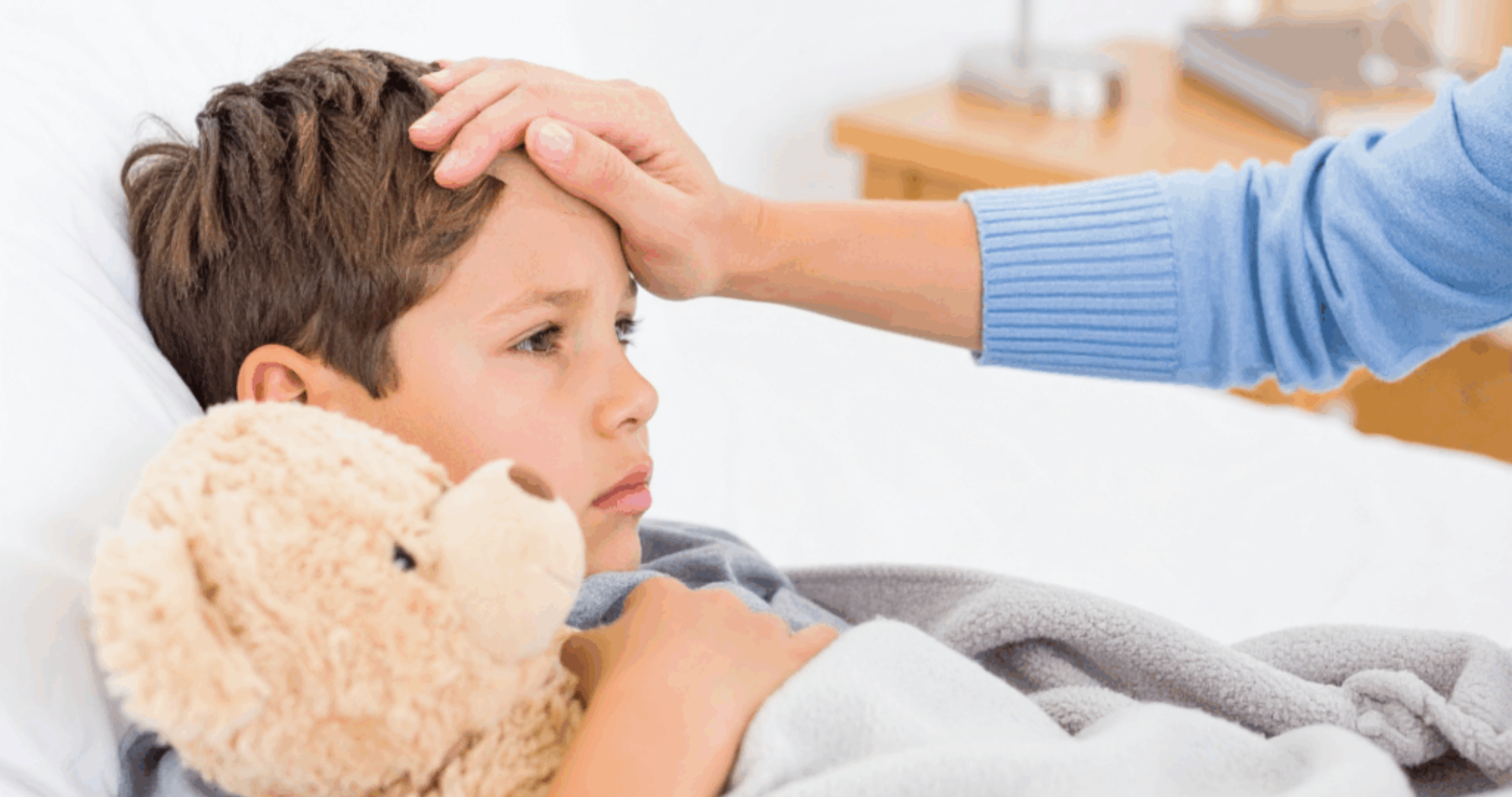 Mother checking child’s forehead temperature while comforting them with a teddy bear.